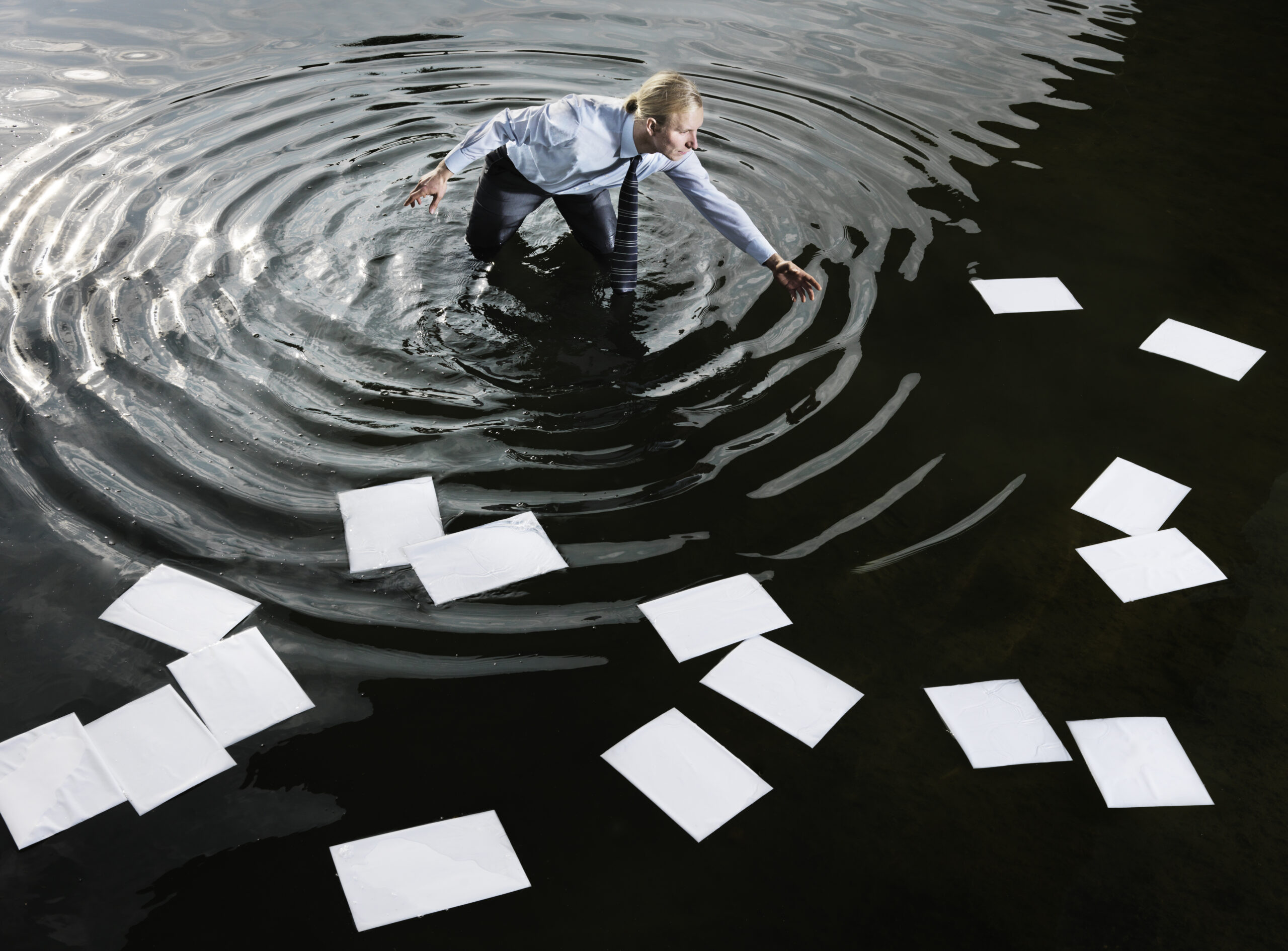 Businessman standing in water collecting floating pieces of A4 paper
